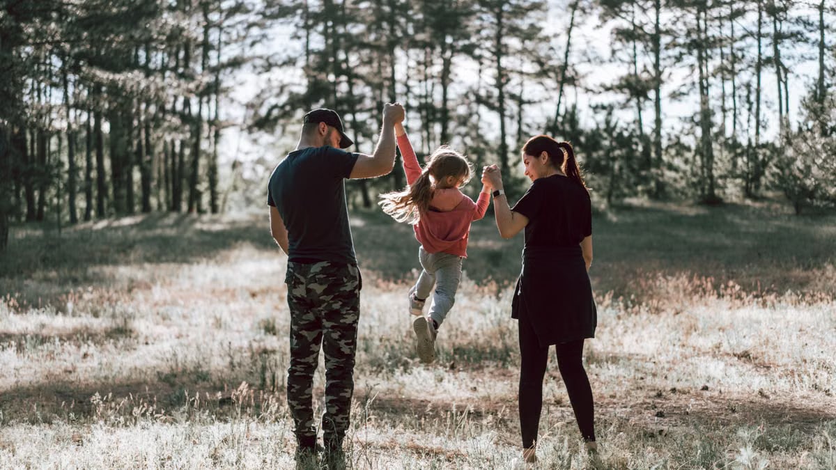 Full body back view of happy parents and daughter in casual clothes holding hands and walking on grassy meadow near forest on sunny summer day